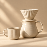 a coordinated cream ceramic pour-over coffee kit: ribbed dripper resting on a carafe, one matching cup beside it, no extra objects